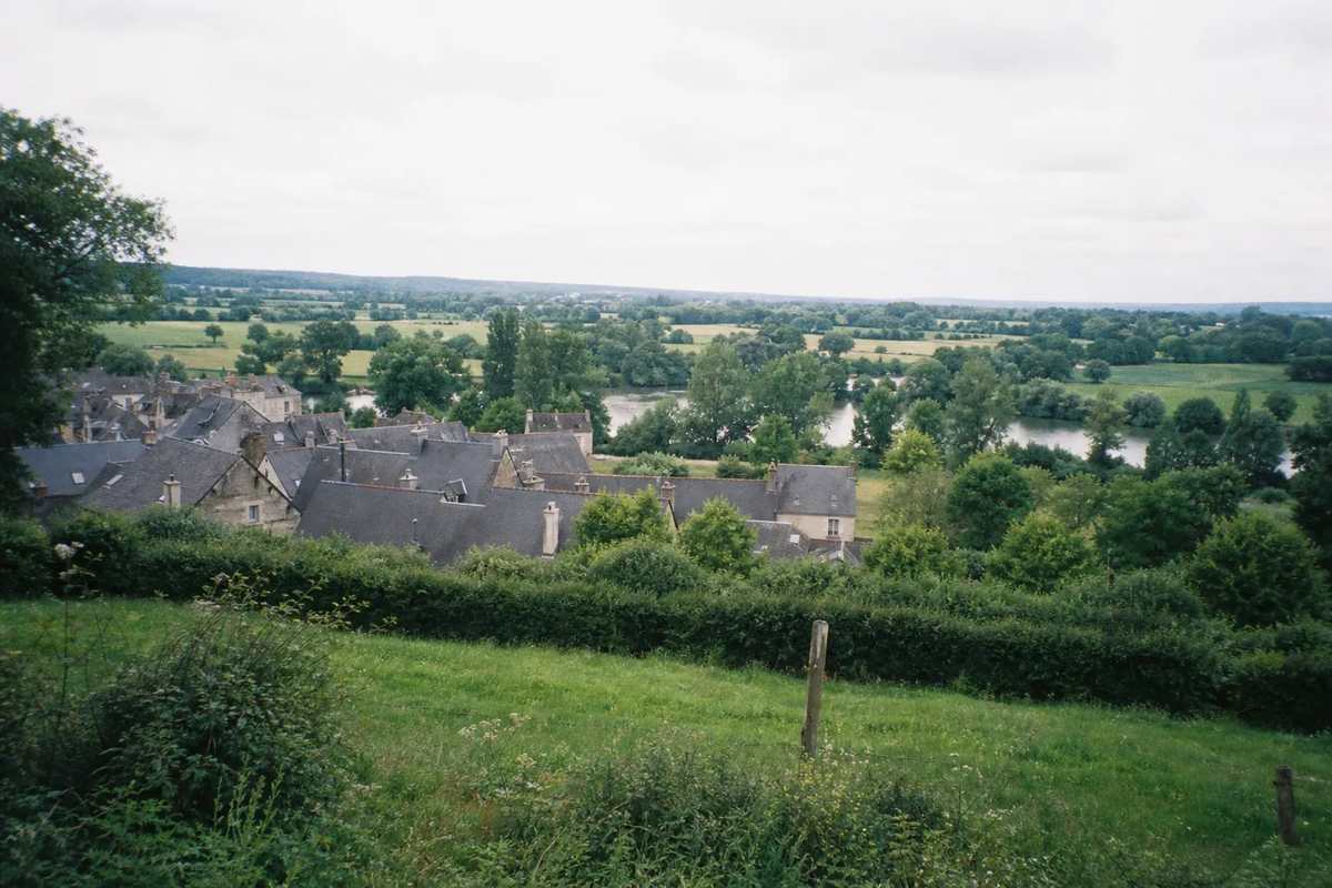 Paysage bocager mayennais où nous intervenons : haies, toits d’ardoise groupés d’un bourg et rivière au loin